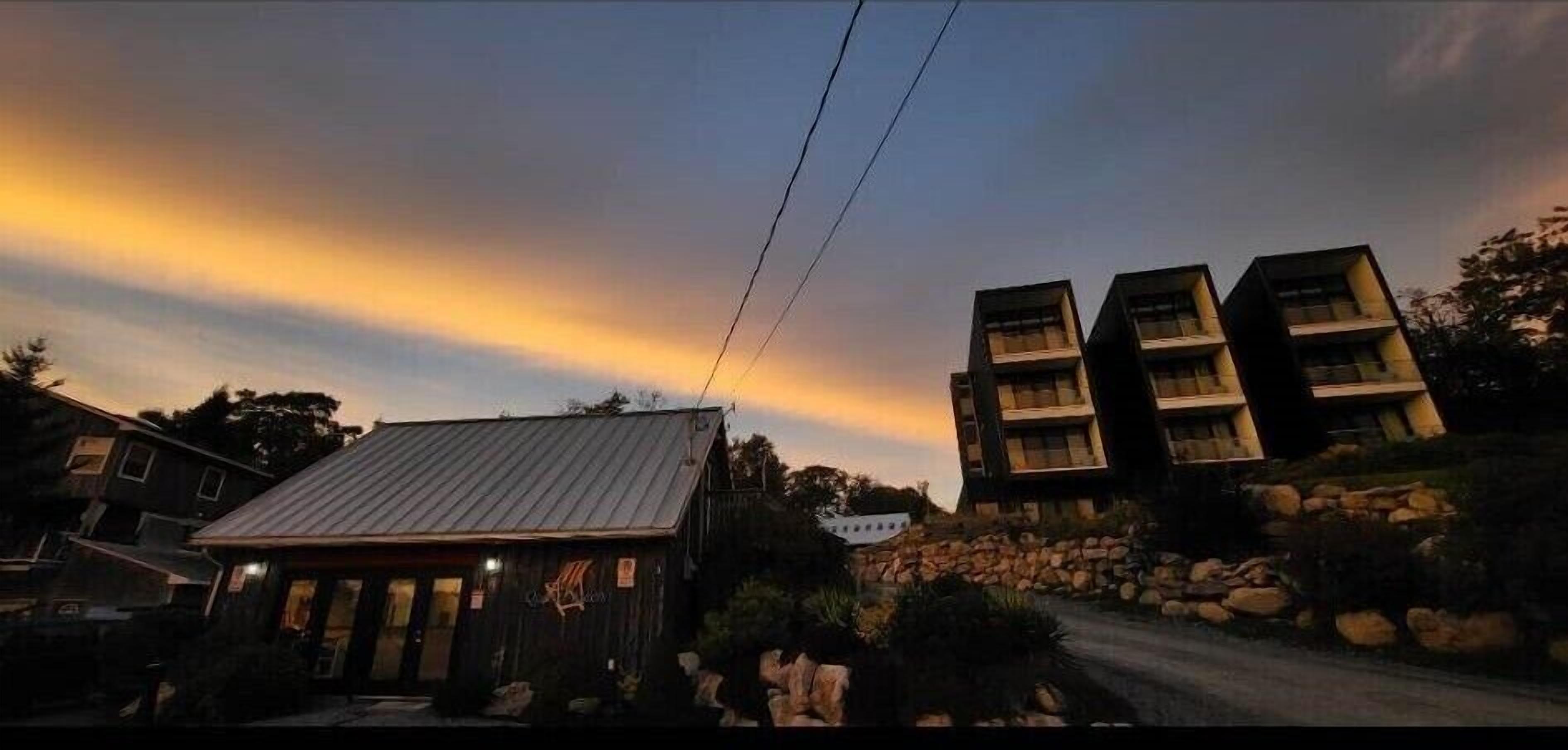 A rustic building with a metal roof and a small porch, adjacent to modern multi-story buildings with balconies.