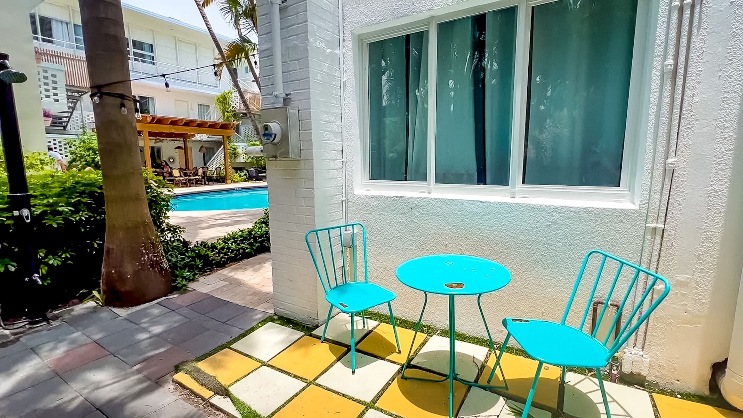 Eine Terrasse mit zwei türkisfarbenen Stühlen und einem kleinen Tisch, im Hintergrund ein Pool und ein weißes Gebäude mit einem Fenster.