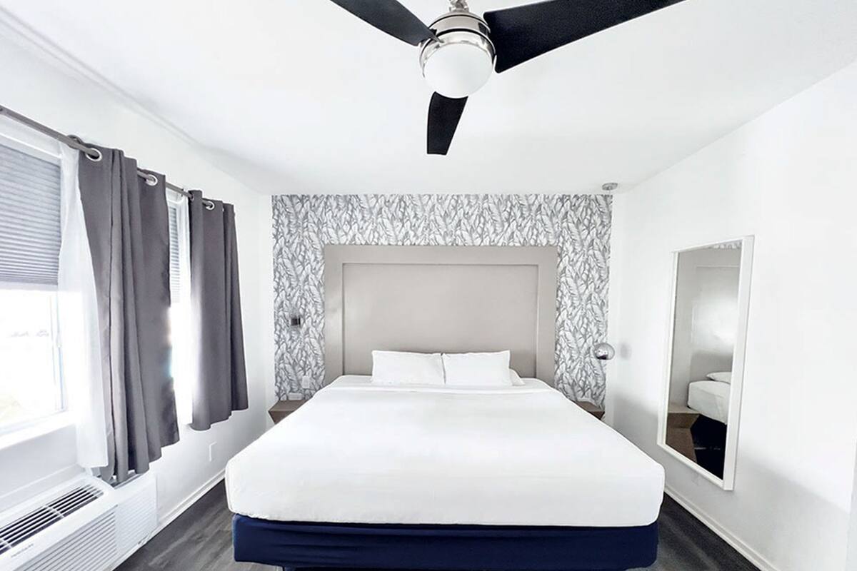 Moderni hotellihuone, jossa on suuri sänky, kattotuuletin, ikkuna verhoilla ja kuvioitu seinä.