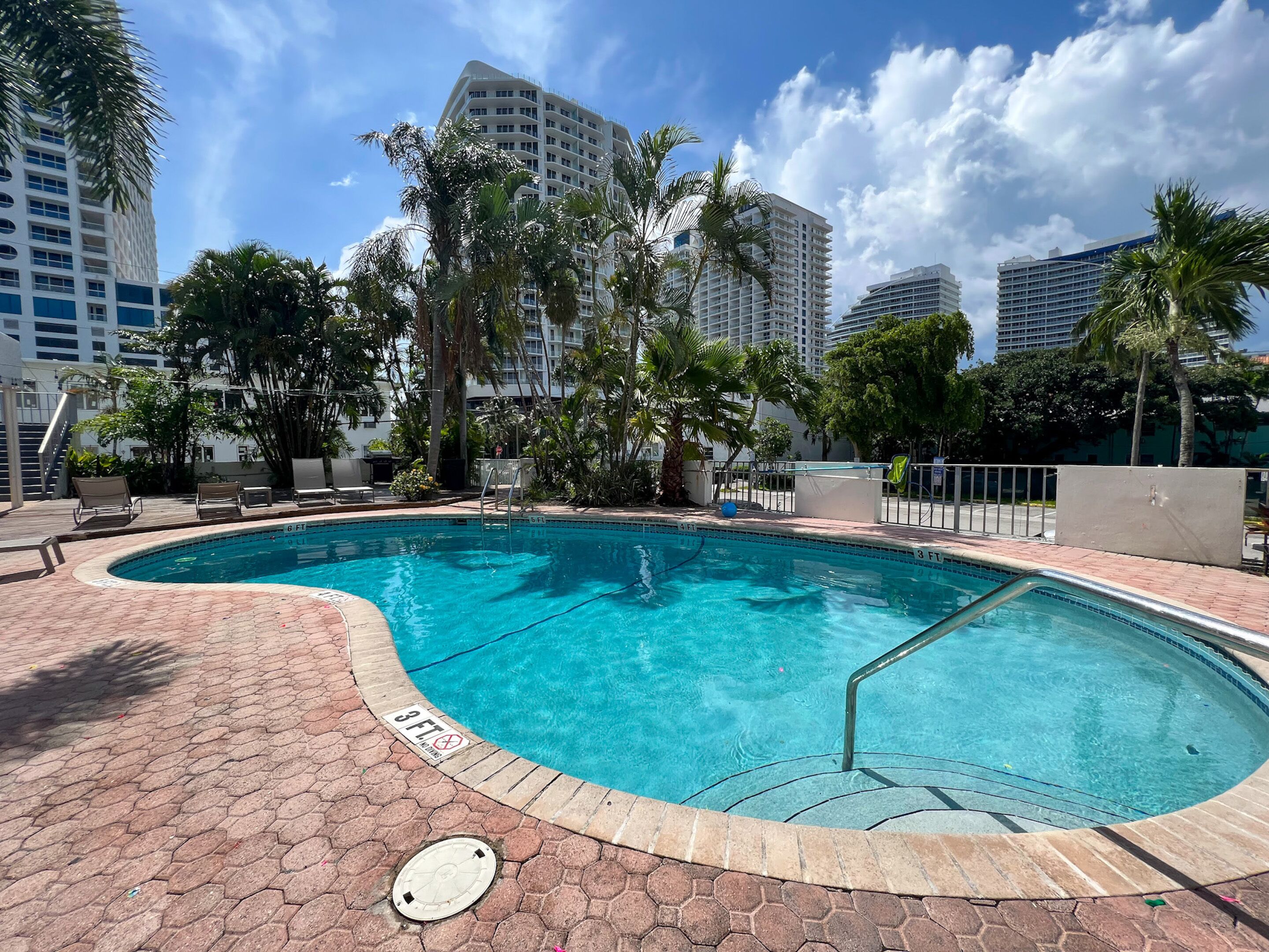 A swimming pool with a 3 FT depth marker, surrounded by palm trees and modern buildings.