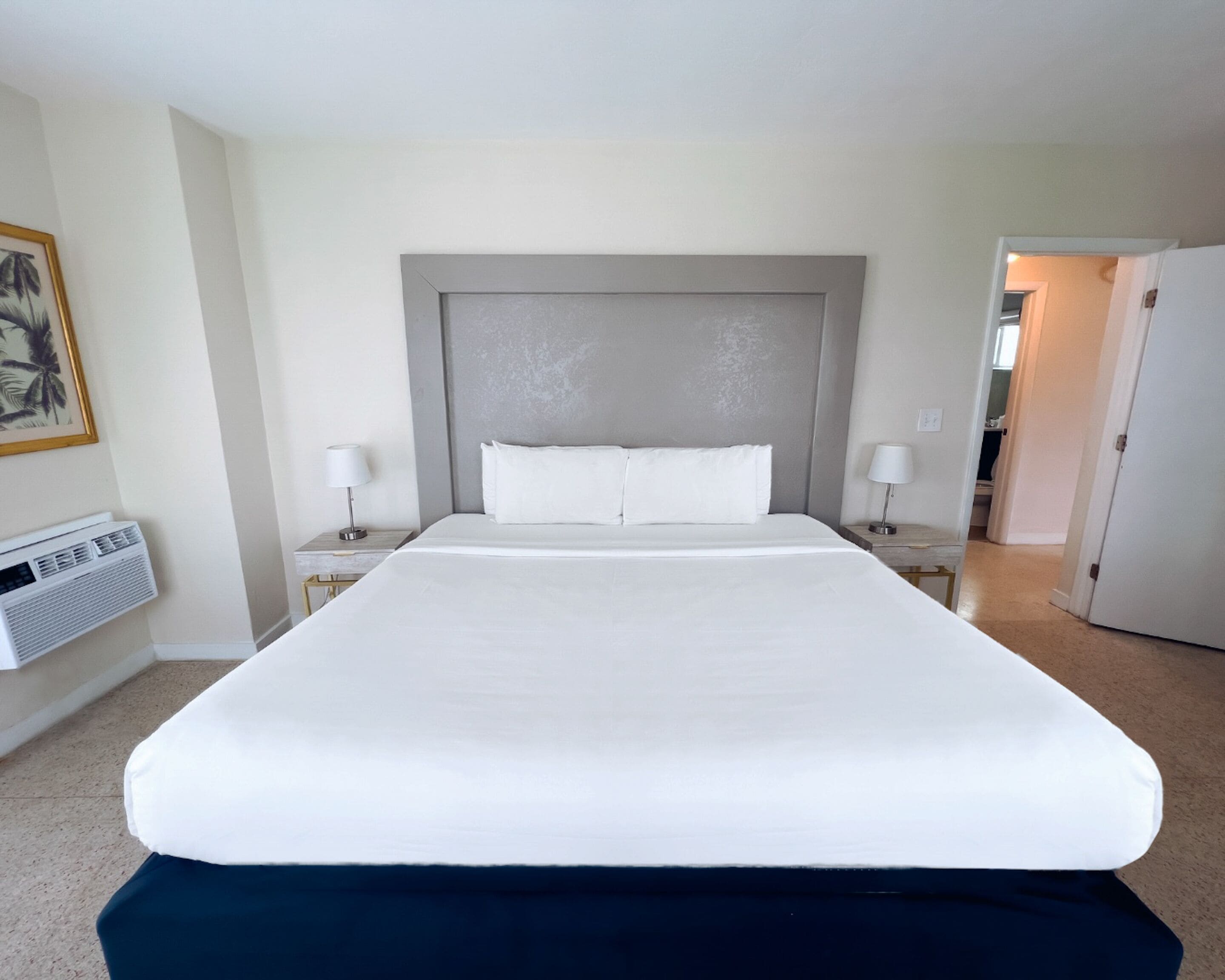 Hotellihuone, jossa on suuri sänky, kaksi yöpöytää, seinälle asennettu ilmastointilaite ja ovi, joka johtaa toiseen huoneeseen.