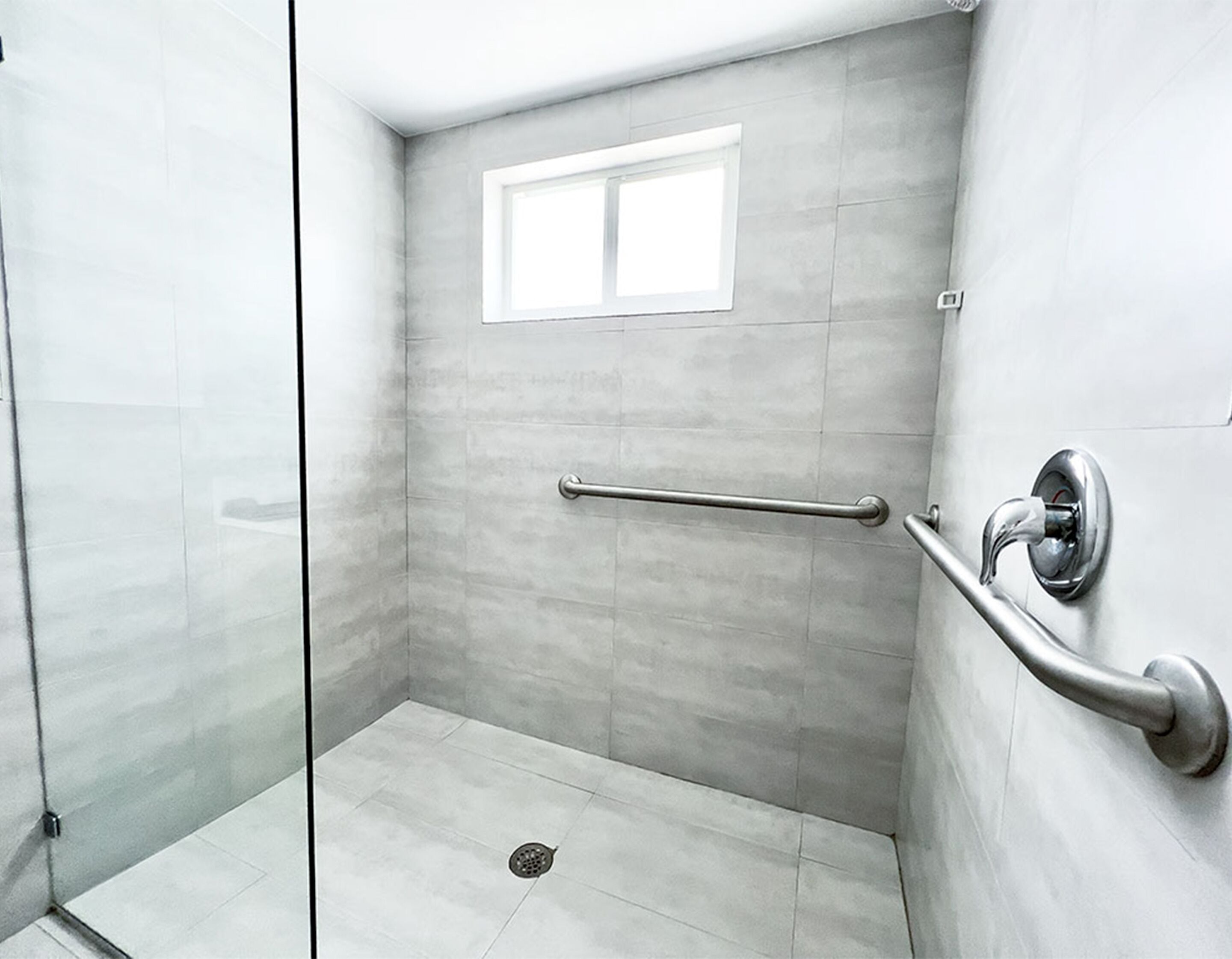 A modern shower area with a glass door, a handrail, a window, and a tiled floor.