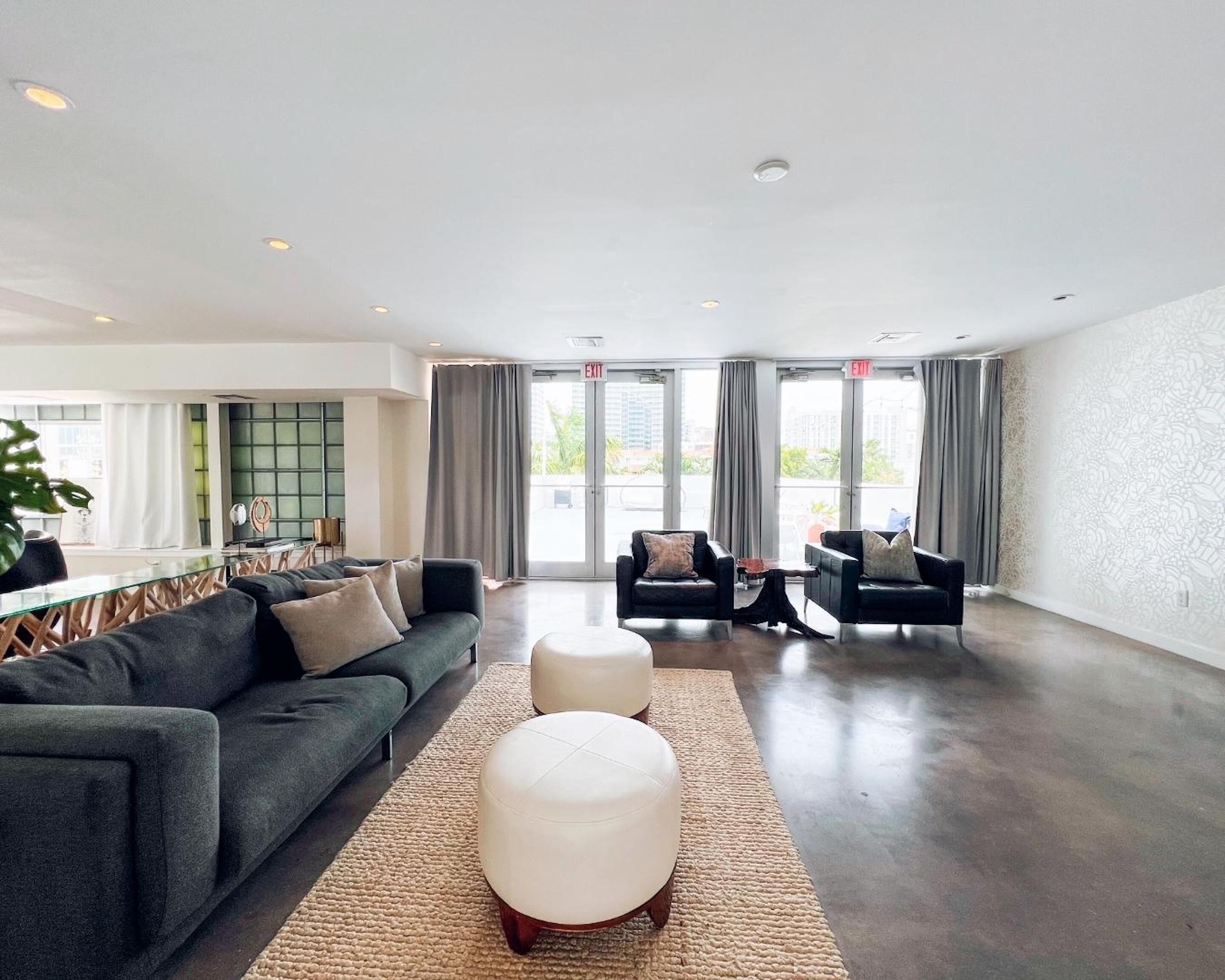 A spacious living area with a sofa set, ottoman, and chairs, a glass coffee table, and a view through large windows.