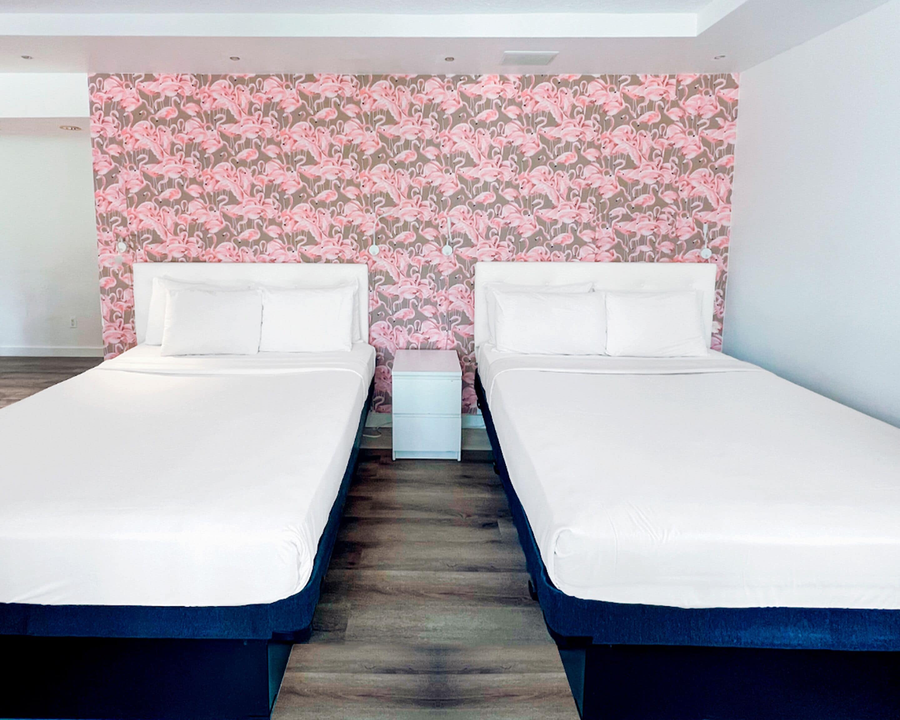 Una habitación de hotel con dos camas individuales, suelo de madera y papel tapiz con flamencos rosas.
