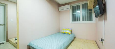 A small, single-bed room with a wooden floor, a light blue bedspread, a window with blinds, an air conditioning unit, and a mounted television.