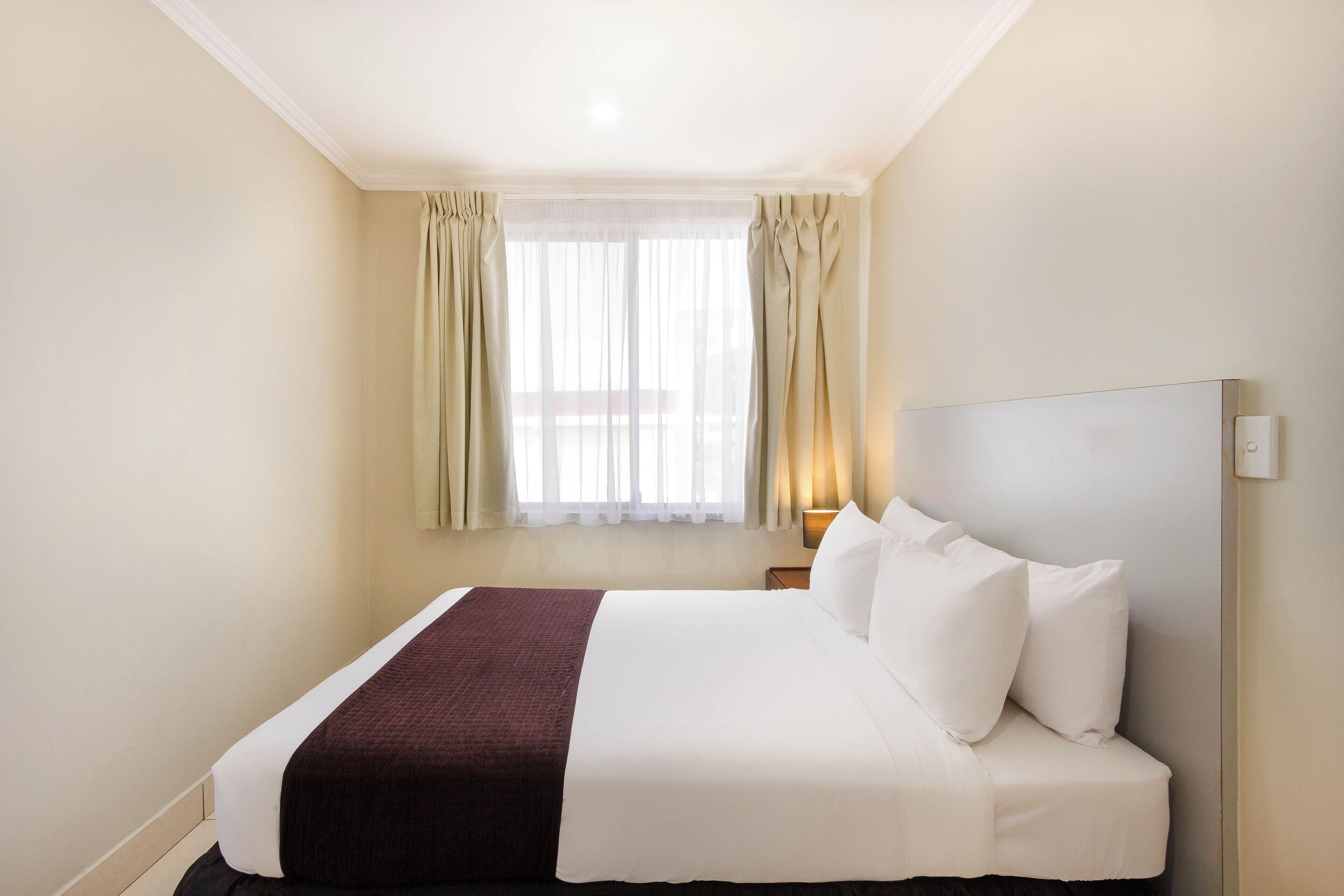Ein Hotelzimmer mit einem aufgeräumten Bett, einem Nachttisch mit Lampe und einem Fenster mit Vorhängen.