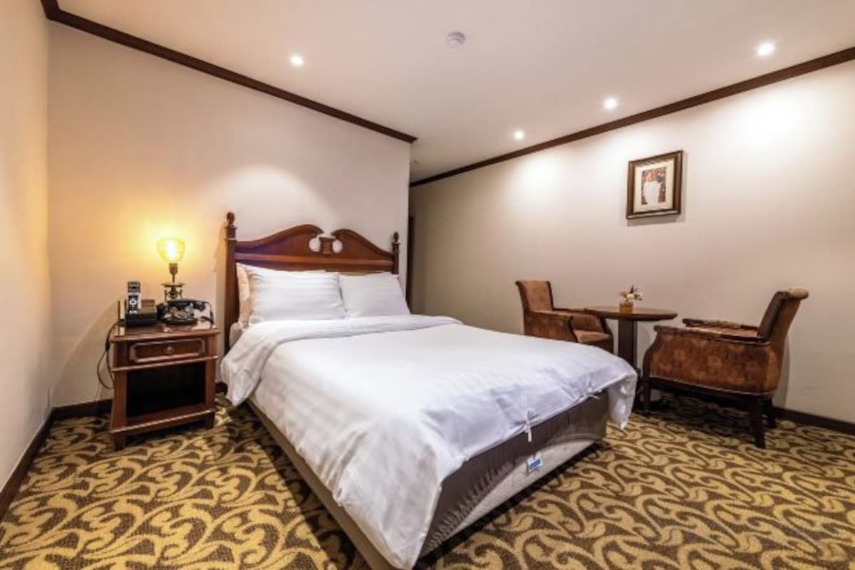 A hotel room with a double bed, a bedside table with a lamp, a chair, and a small table.