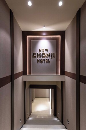 A modern hotel interior with a central sign reading 'NEW CHONJI HOTEL,' a staircase leading to the upper floor, and a clean, minimalist design.