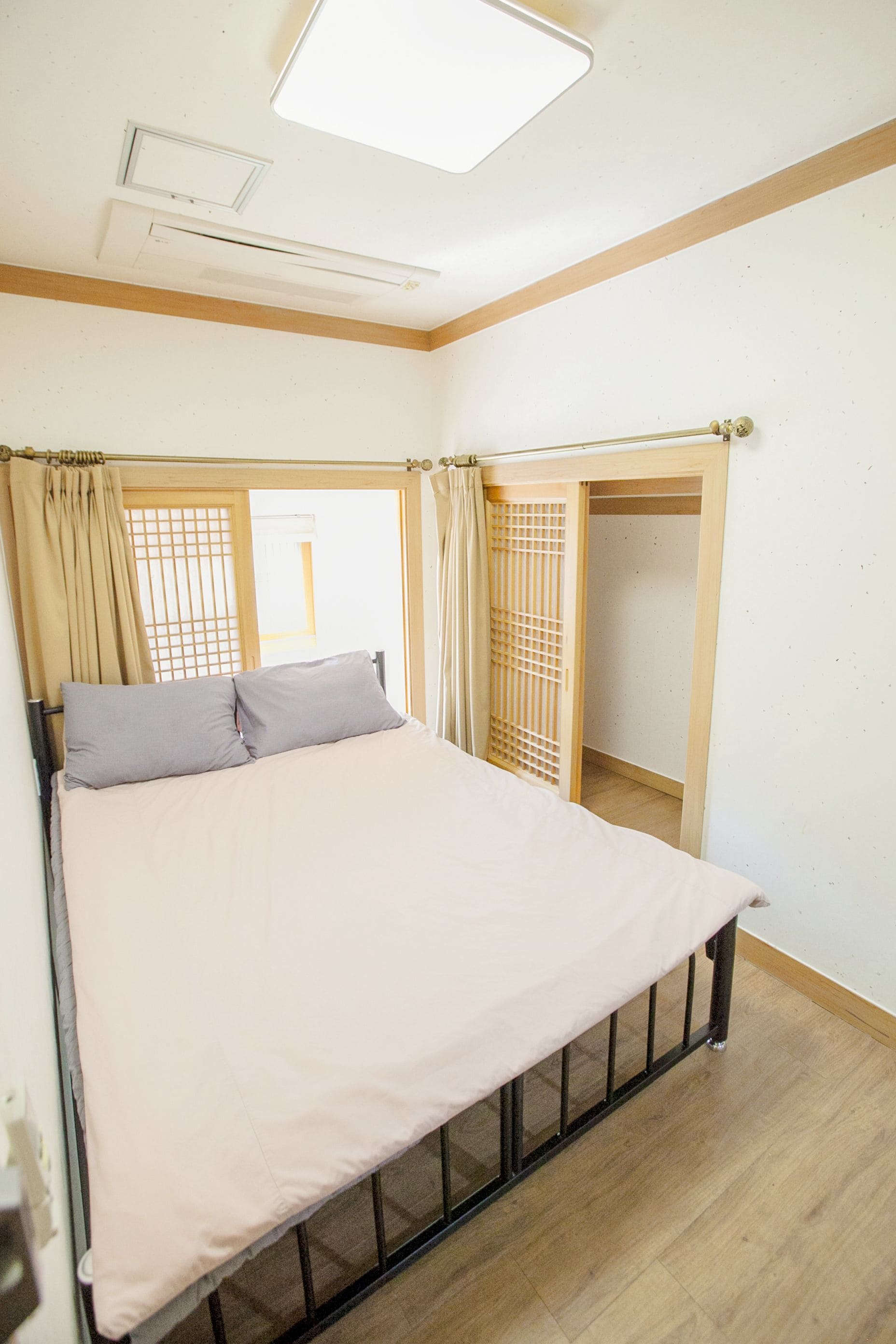 A bedroom with a bed, a window with curtains, a wooden floor, and a ceiling light.