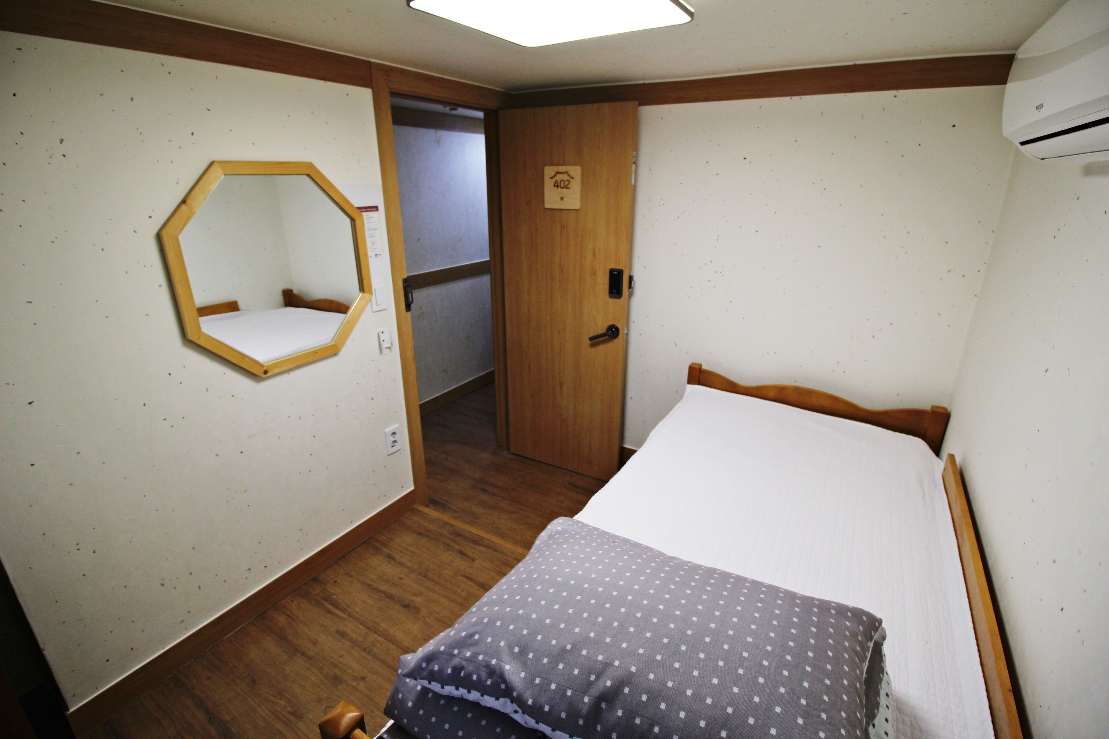 A small, compact room with a wooden floor, a bed with a white mattress and a grey polka dot blanket, a wooden-framed mirror on the wall, and a door with a number sign.