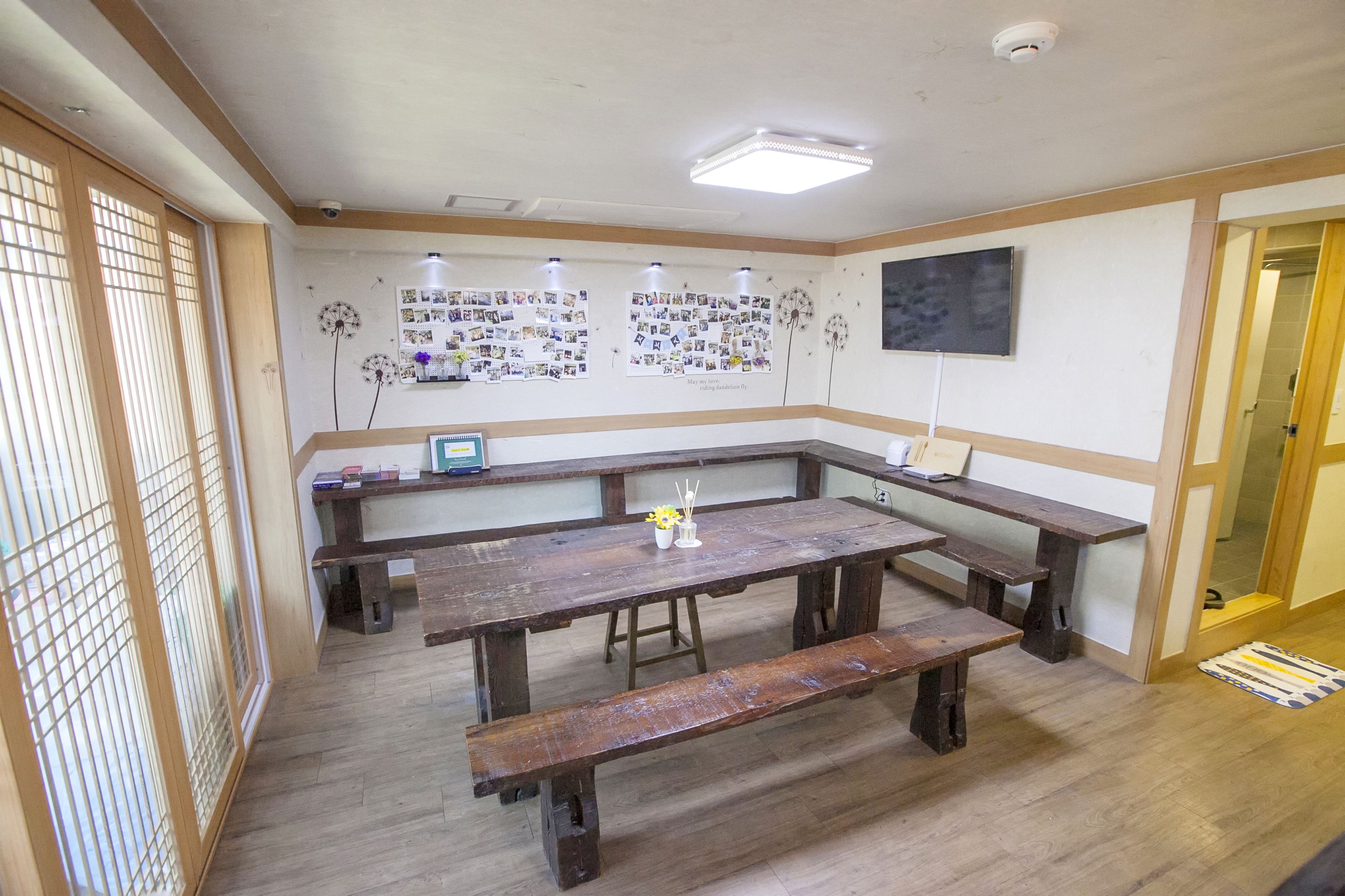 A room with wooden benches and a table, a bulletin board with photos, a flat-screen TV, and a ceiling-mounted security camera.