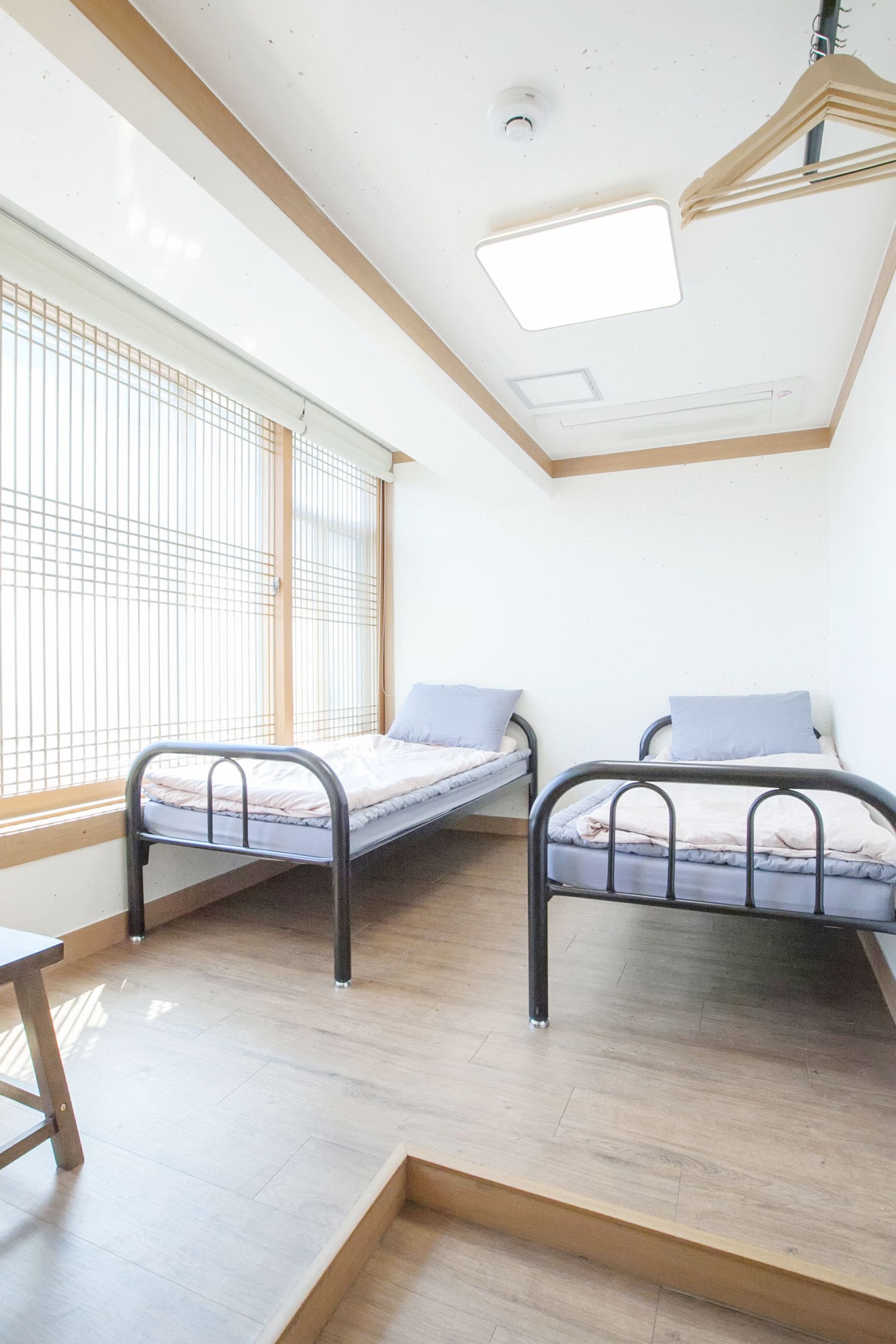 A room with two metal bunk beds, a wooden floor, a window with blinds, and a ceiling light fixture.