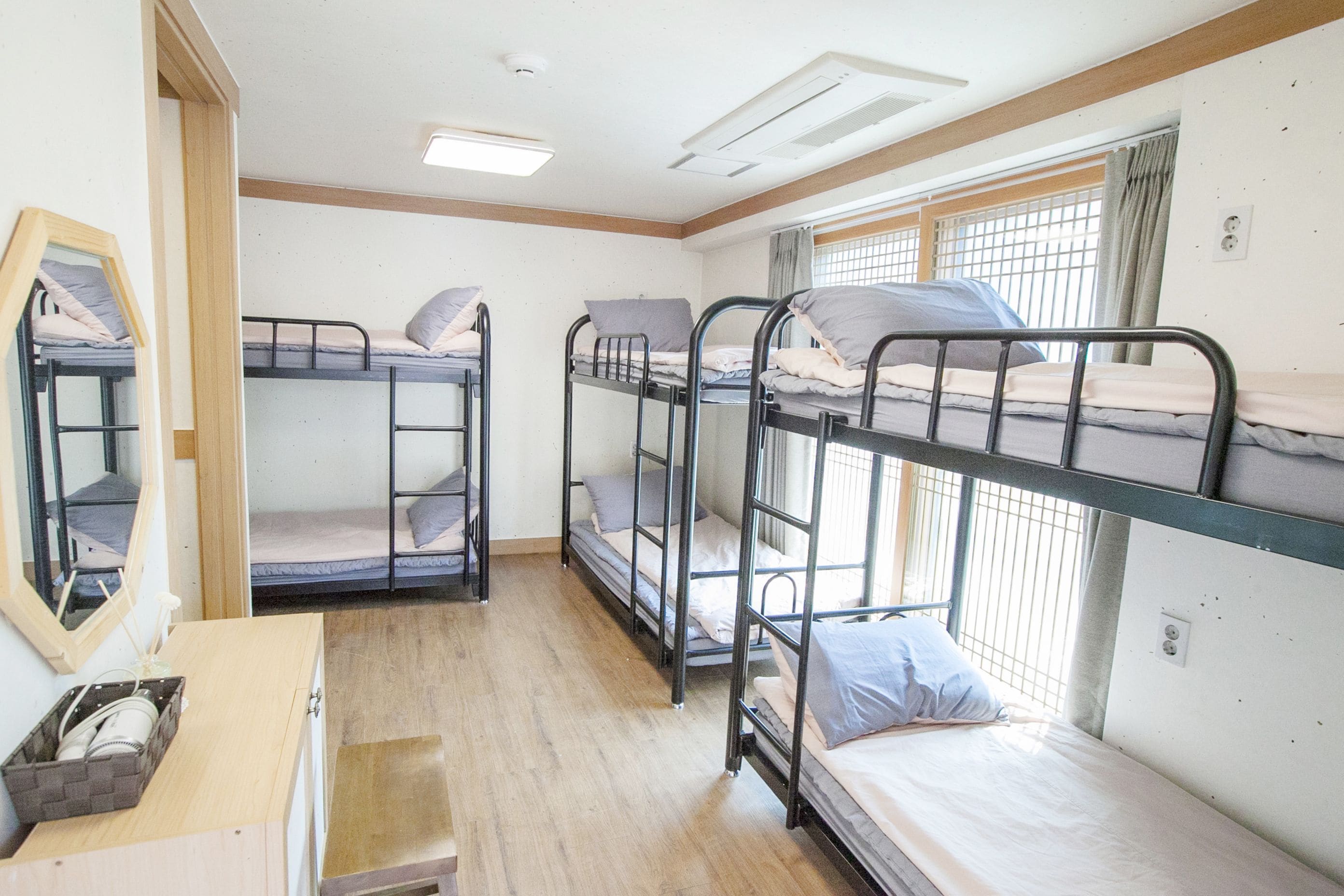 A dormitory room with bunk beds, a wooden desk, a mirror, and a window with curtains.