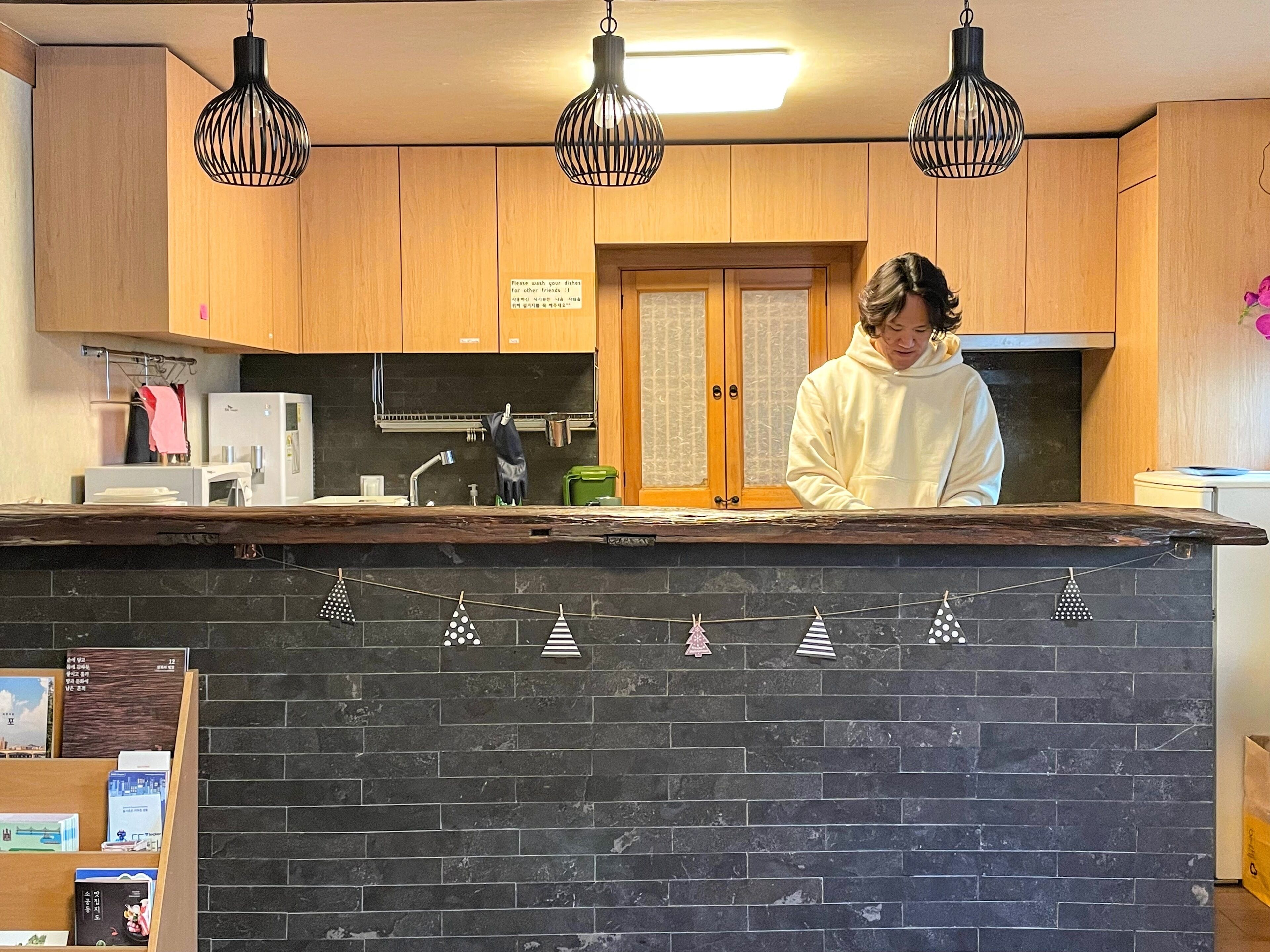 Une cuisine avec des meubles en bois, un plan de travail noir, des luminaires suspendus et une personne debout près du comptoir.