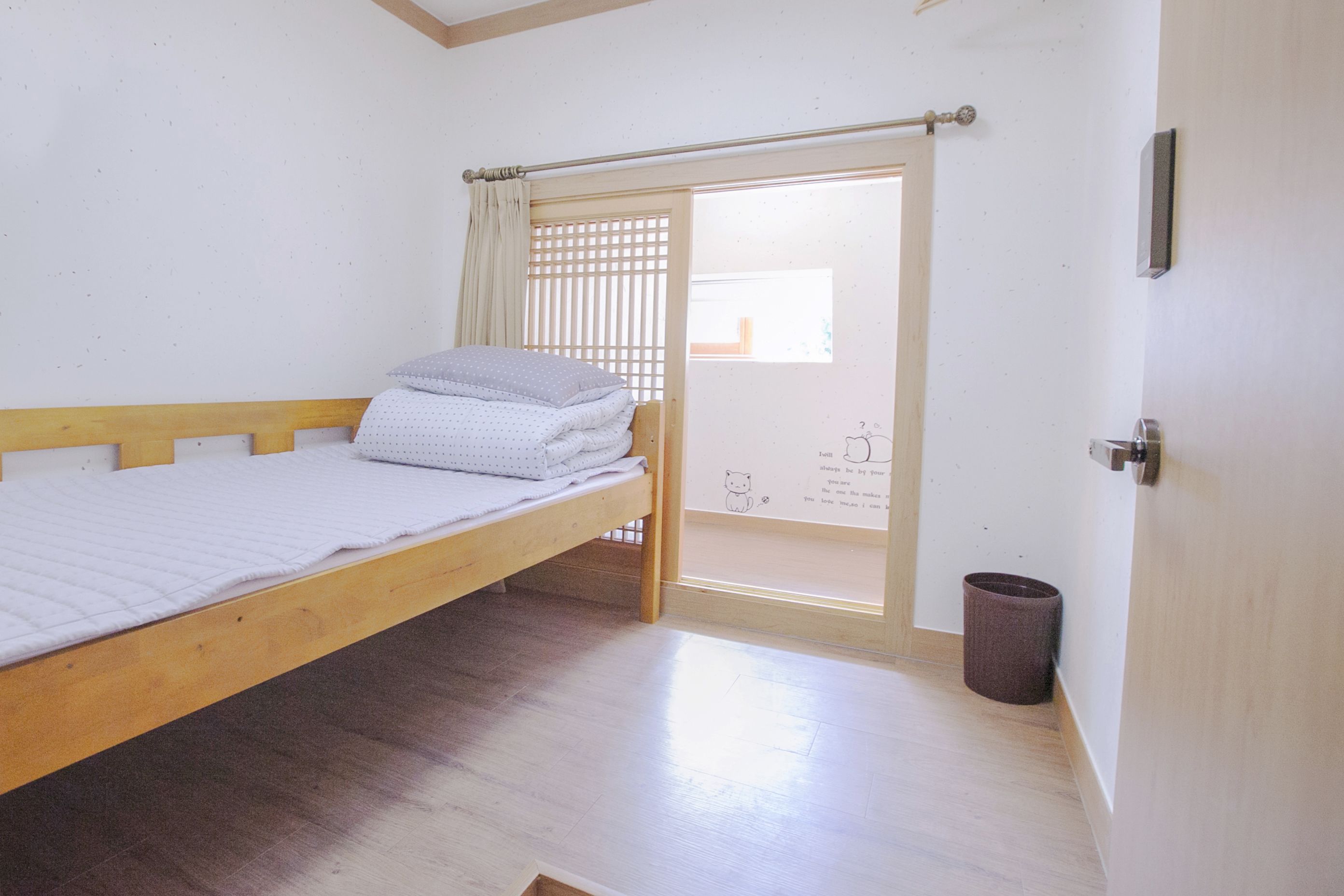 A single bed with a wooden frame and a white mattress, a small trash bin, and a sliding door leading to an outdoor area.