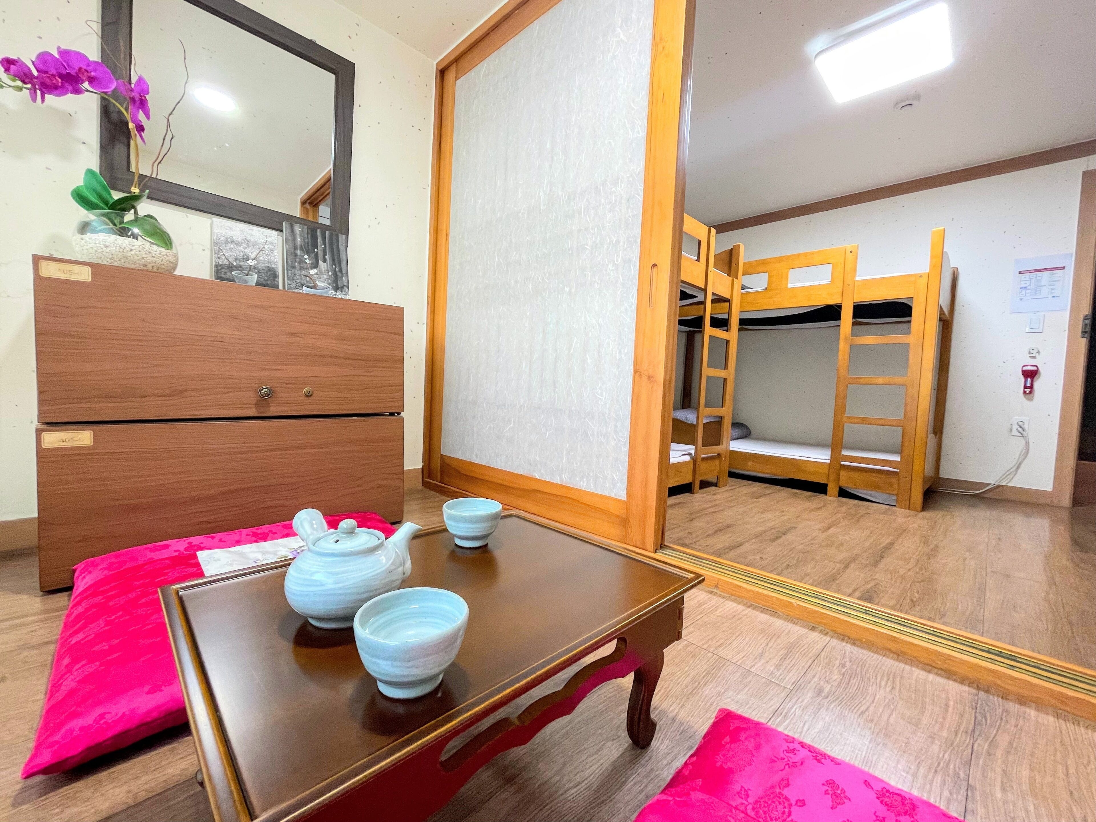 Une pièce comprenant une commode en bois, une table avec une théière et des tasses, un lit superposé et un miroir.