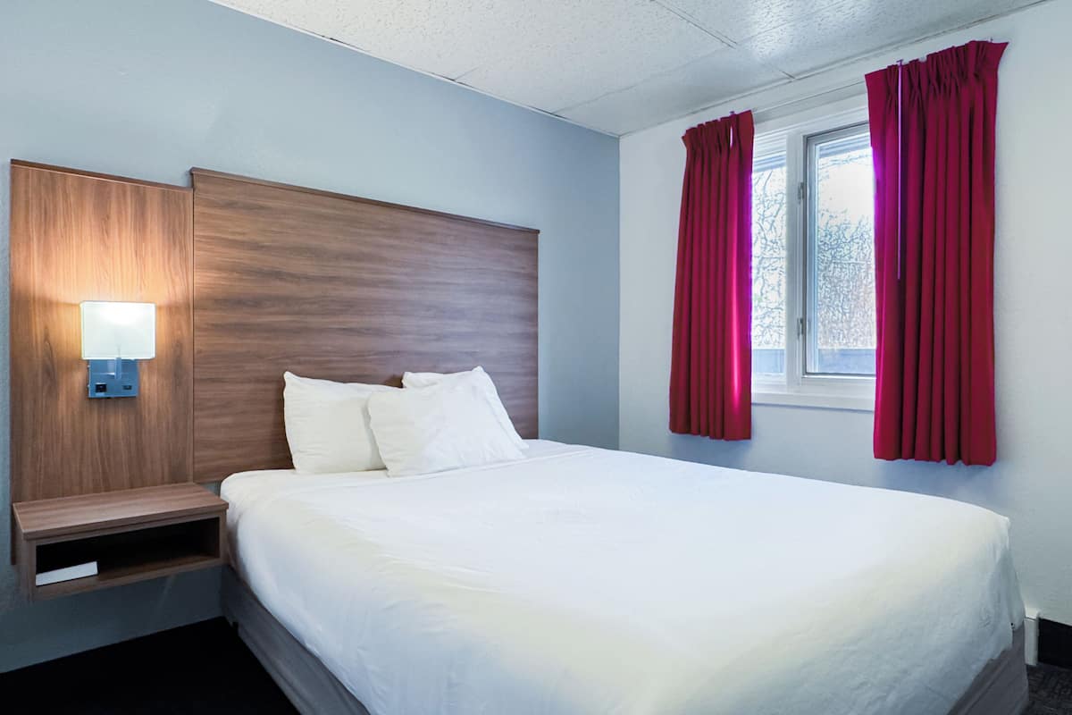 A hotel room with a wooden headboard, a bedside table with a lamp, a large bed with white linens, and red curtains on the window.