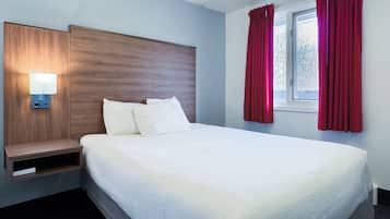 A hotel room with a wooden headboard, a bedside table with a lamp, a large bed with white linens, and red curtains on the window.