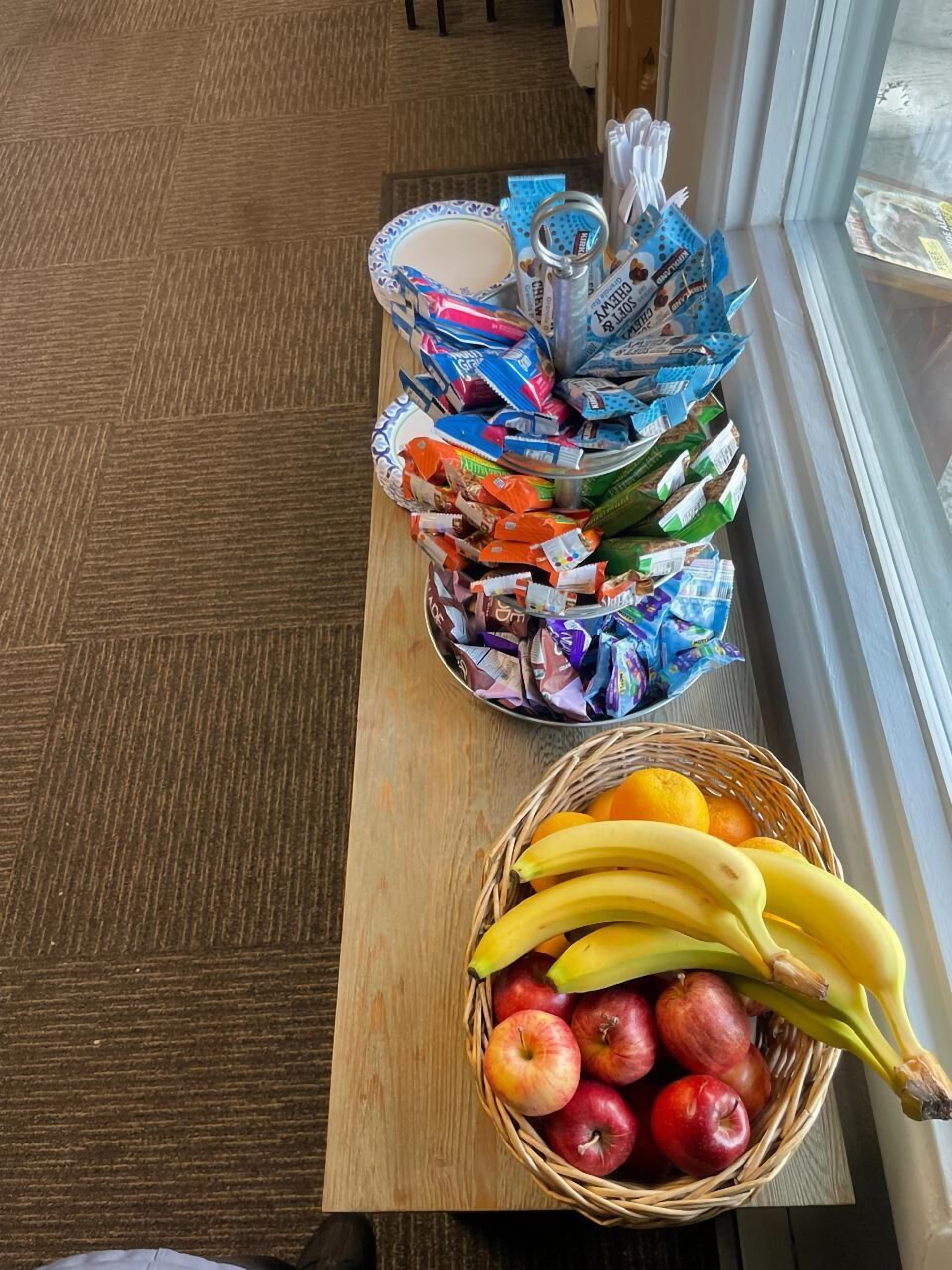 A wooden table with a basket of apples and bananas, and a rack of snack packets.
