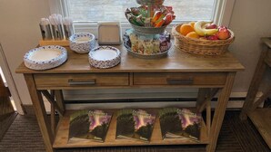 A wooden table with a window view, containing plates, bowls, a fruit basket, and a tray of snacks. - Golden North Inn (Fairbanks)