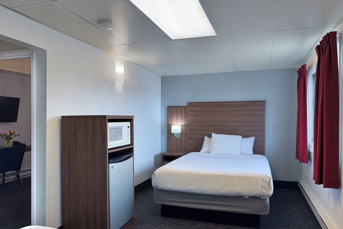 A hotel room with a bed, a desk with a microwave, a TV, and a small table with flowers.