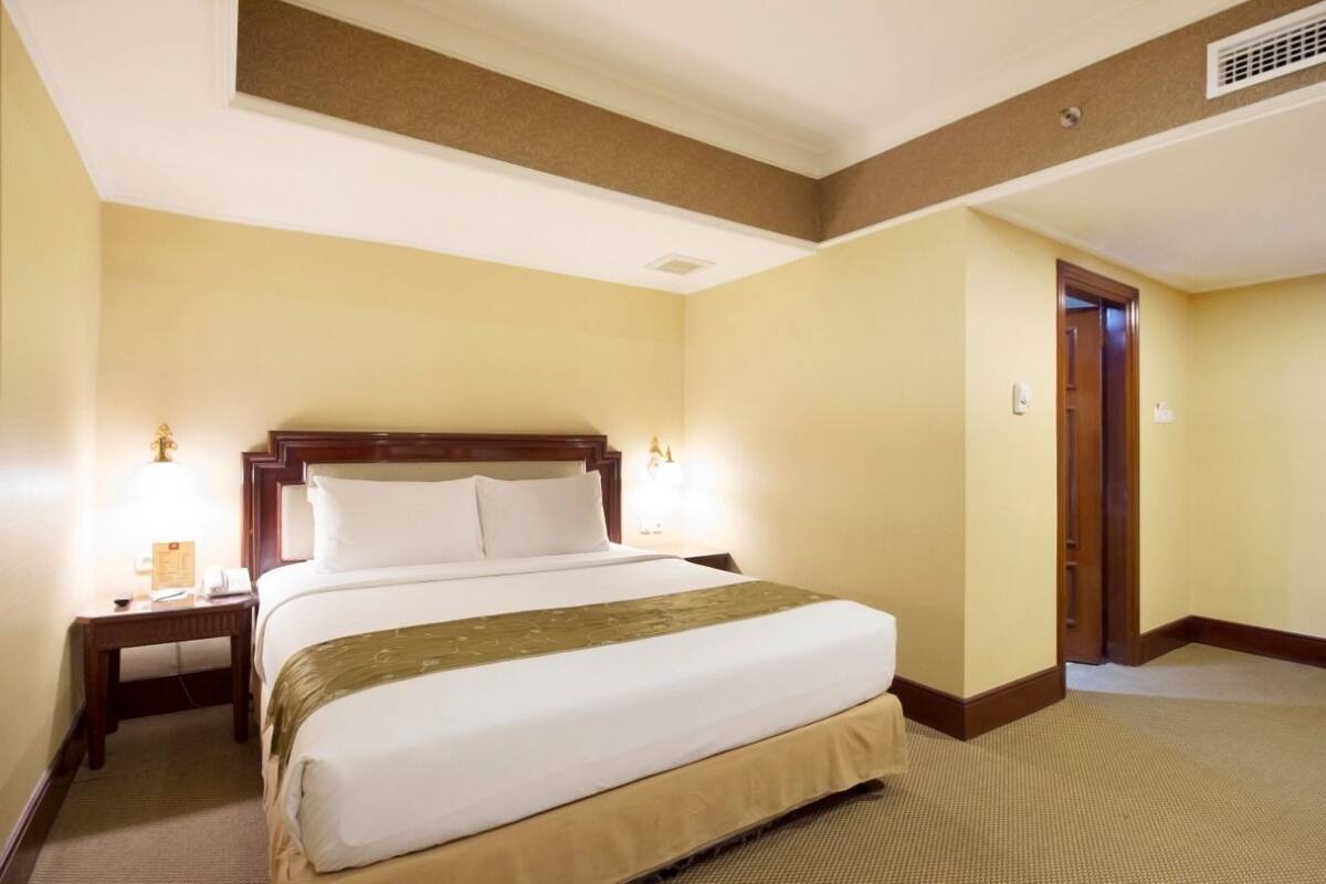 A hotel room with a large bed, bedside lamps, a wooden headboard, and a carpeted floor.