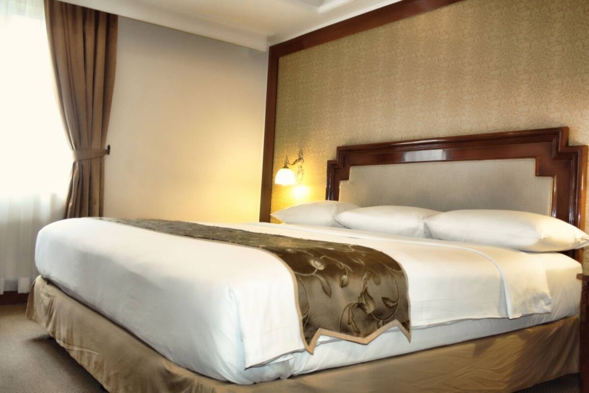A neatly made bed with a headboard, a bedside lamp, and a window with curtains.