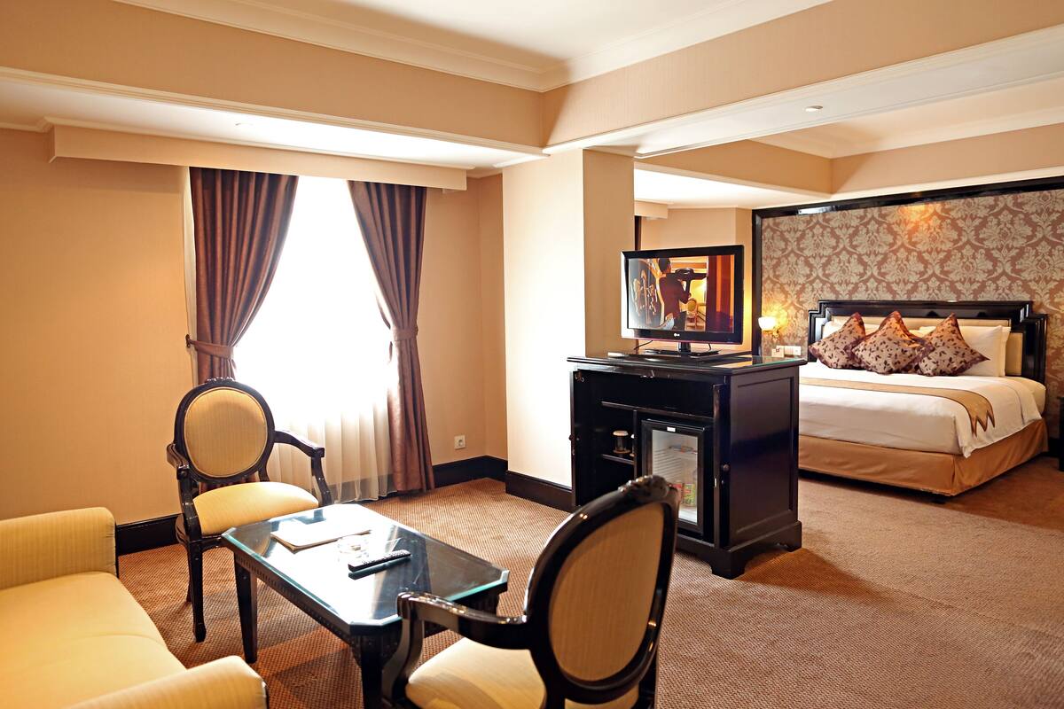 A hotel room with a bed, a desk with a television, two armchairs, and a carpeted floor.