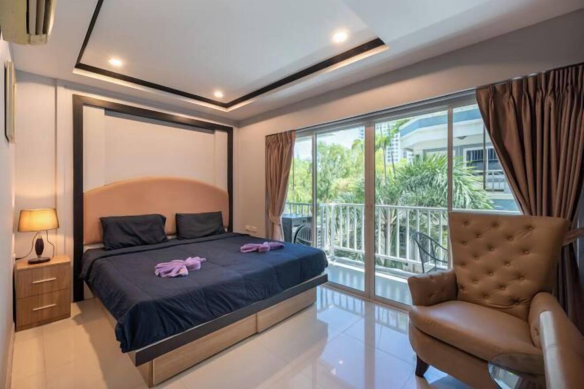 A modern hotel room with a large bed, a sofa, a balcony with a railing, and a view of greenery outside.