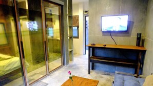 A modern living room with a wooden coffee table, a sofa, a flat-screen TV mounted on the wall, and a sliding glass door leading to another room. - The Cave Studio Samui (Koh Samui)