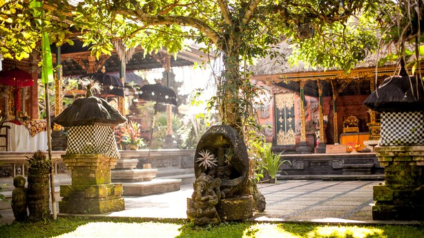 A traditional temple courtyard with stone statues, a checkered lantern, and a thatched-roof structure. - Rumah Desa (Marga)