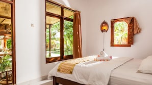 A room with a bed, a window with curtains, a wall-mounted light fixture, and a view of greenery outside. - Rumah Desa (Marga)