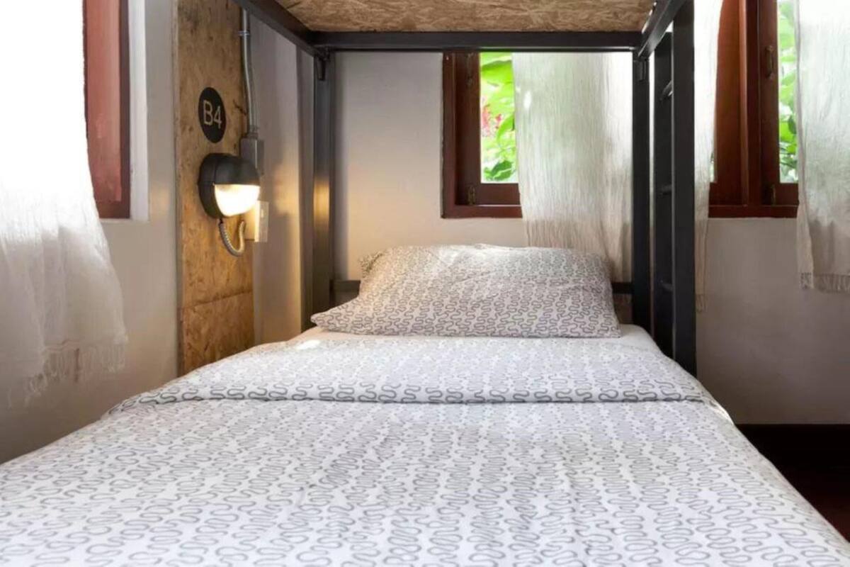 A single bed with a patterned bedspread in a room with a wooden wall, a small window, and a wall-mounted light.