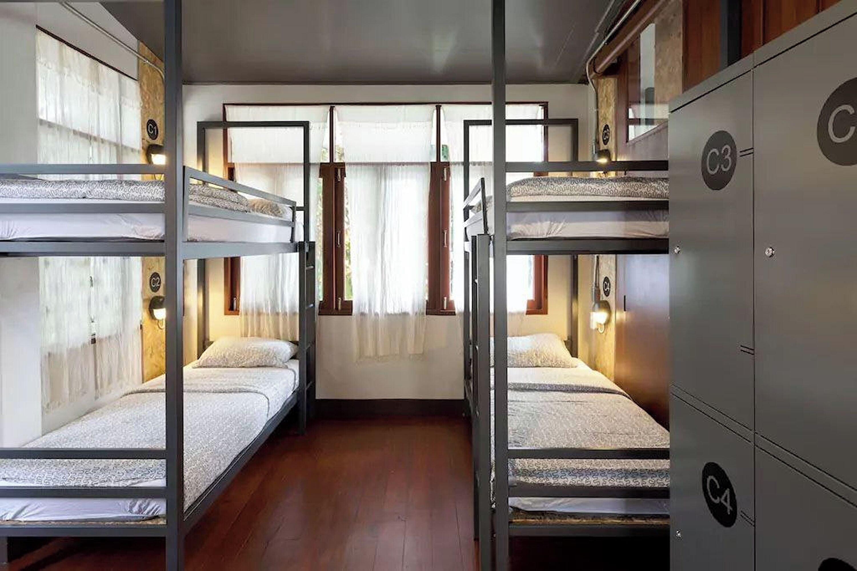 A dormitory room with bunk beds, wooden flooring, and a window with curtains.