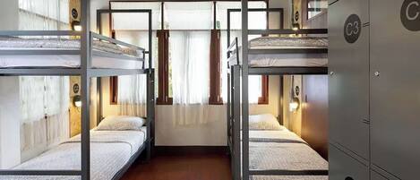 A dormitory room with bunk beds, wooden flooring, and a window with curtains.