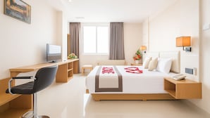 Habitación de hotel con una cama, un escritorio con silla, televisor, mesita de noche con lámpara y ventana con cortinas.