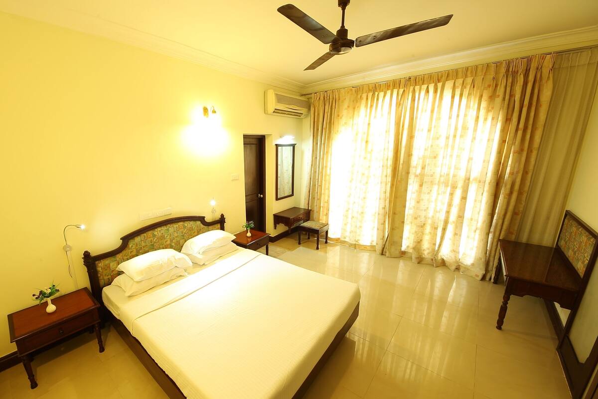 A hotel room with a large bed, a ceiling fan, a desk, a chair, and a window with curtains.