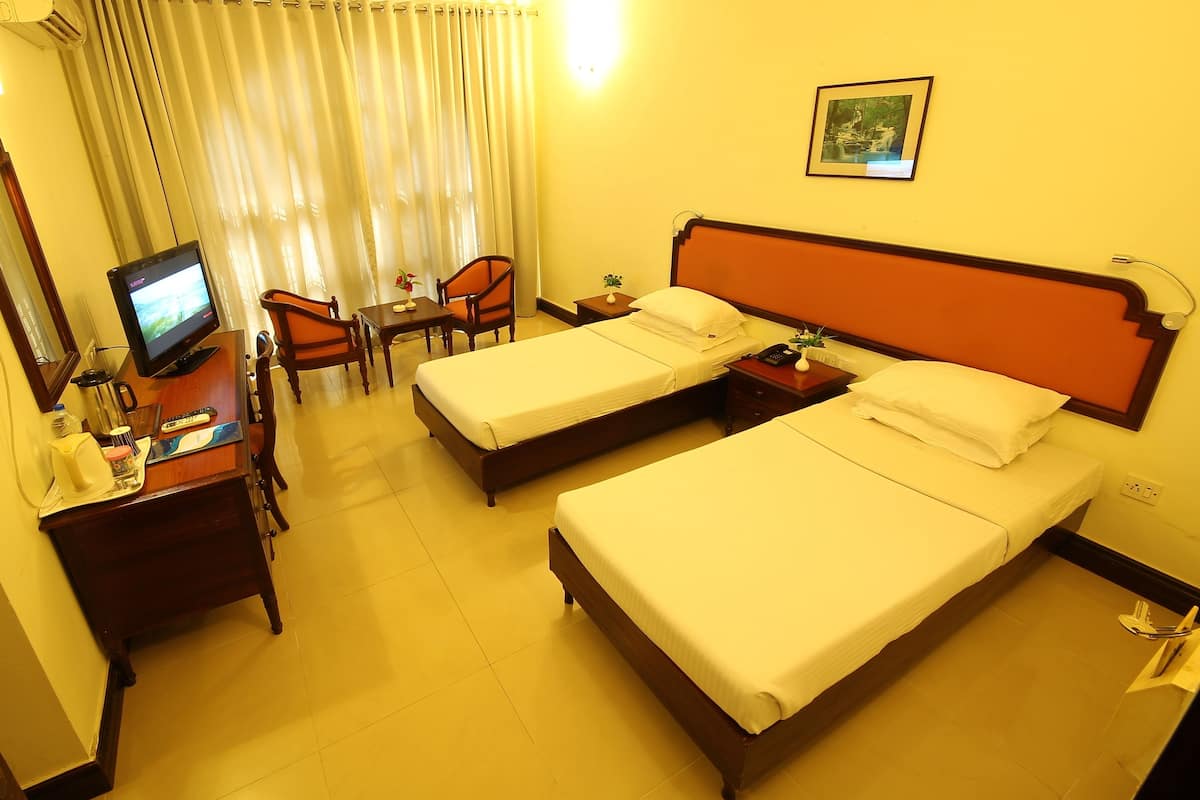 A hotel room with two beds, a desk with a computer, a chair, a small table with a vase, and a ceiling fan.