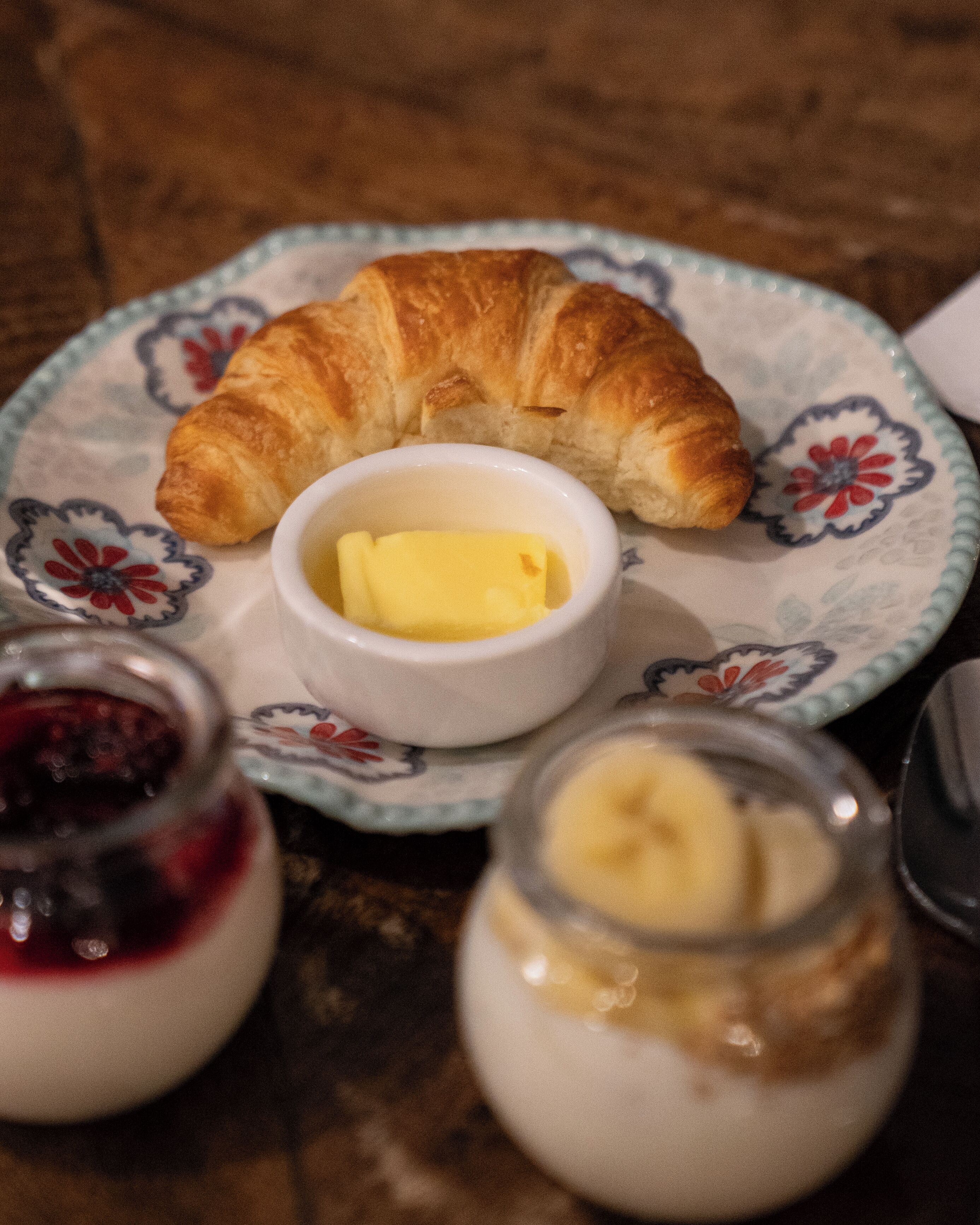 A plate with a croissant and a small bowl of butter, along with two jars of jam and a jar of cream.