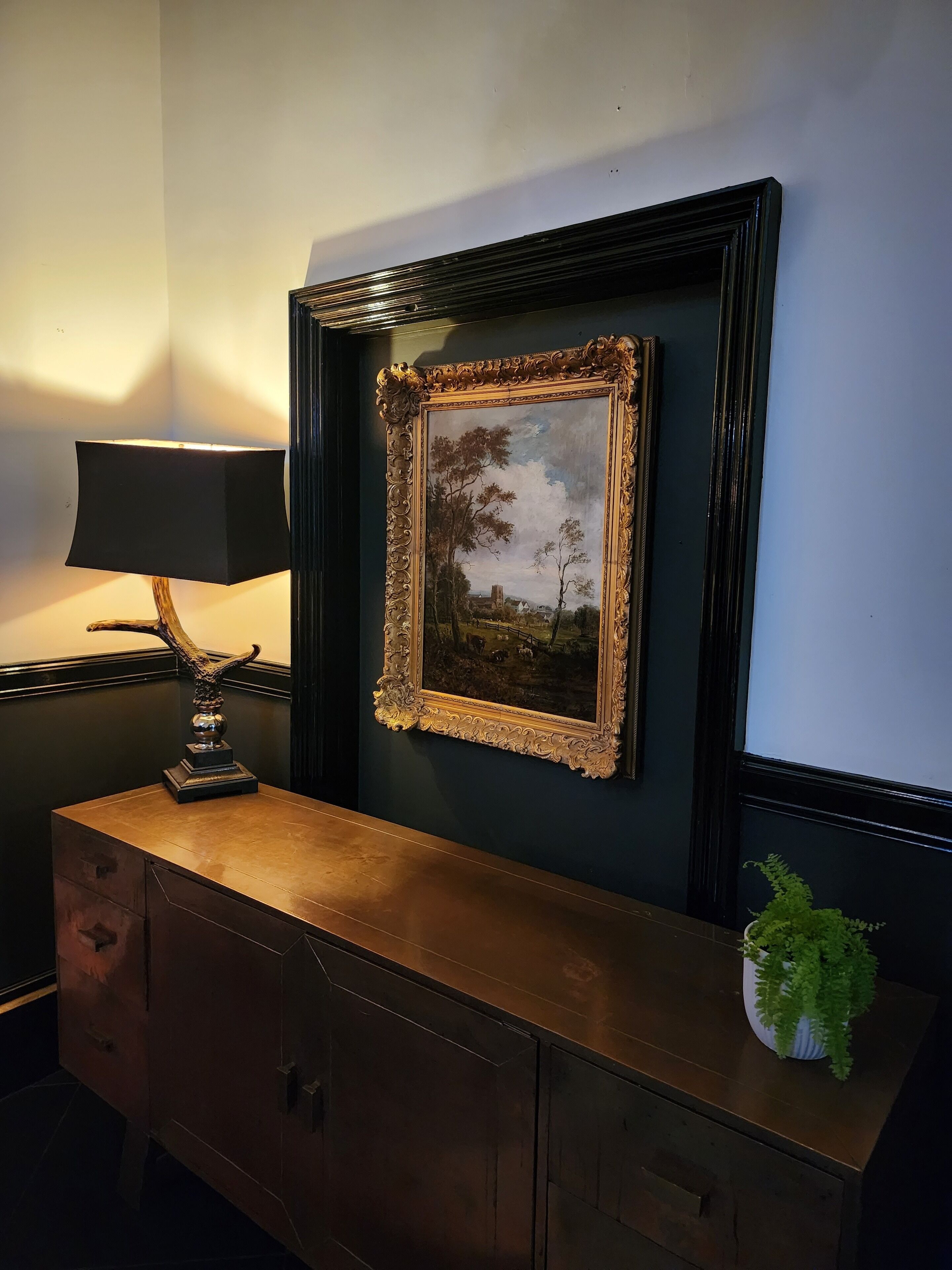 A framed landscape painting on a wall, a lamp, and a cabinet with a plant.
