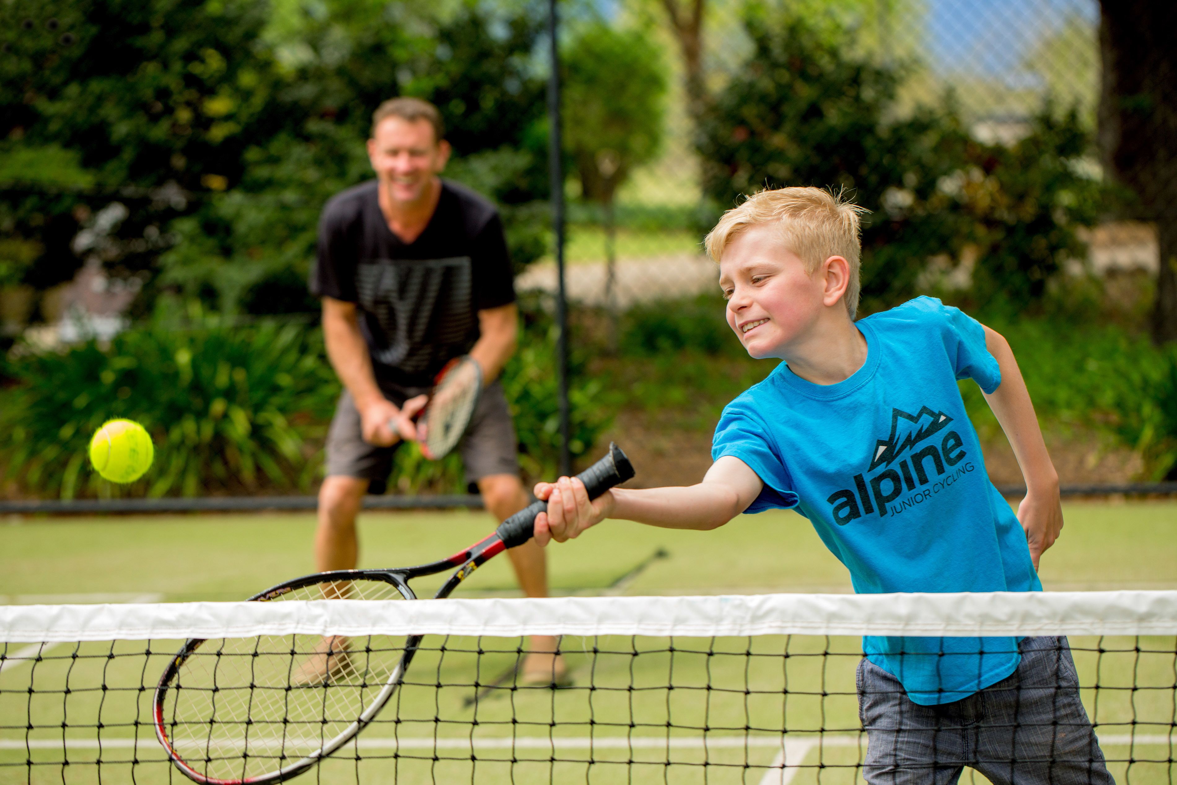 Un court de tennis avec un filet au premier plan et un joueur portant un maillot bleu qui frappe une balle de tennis.