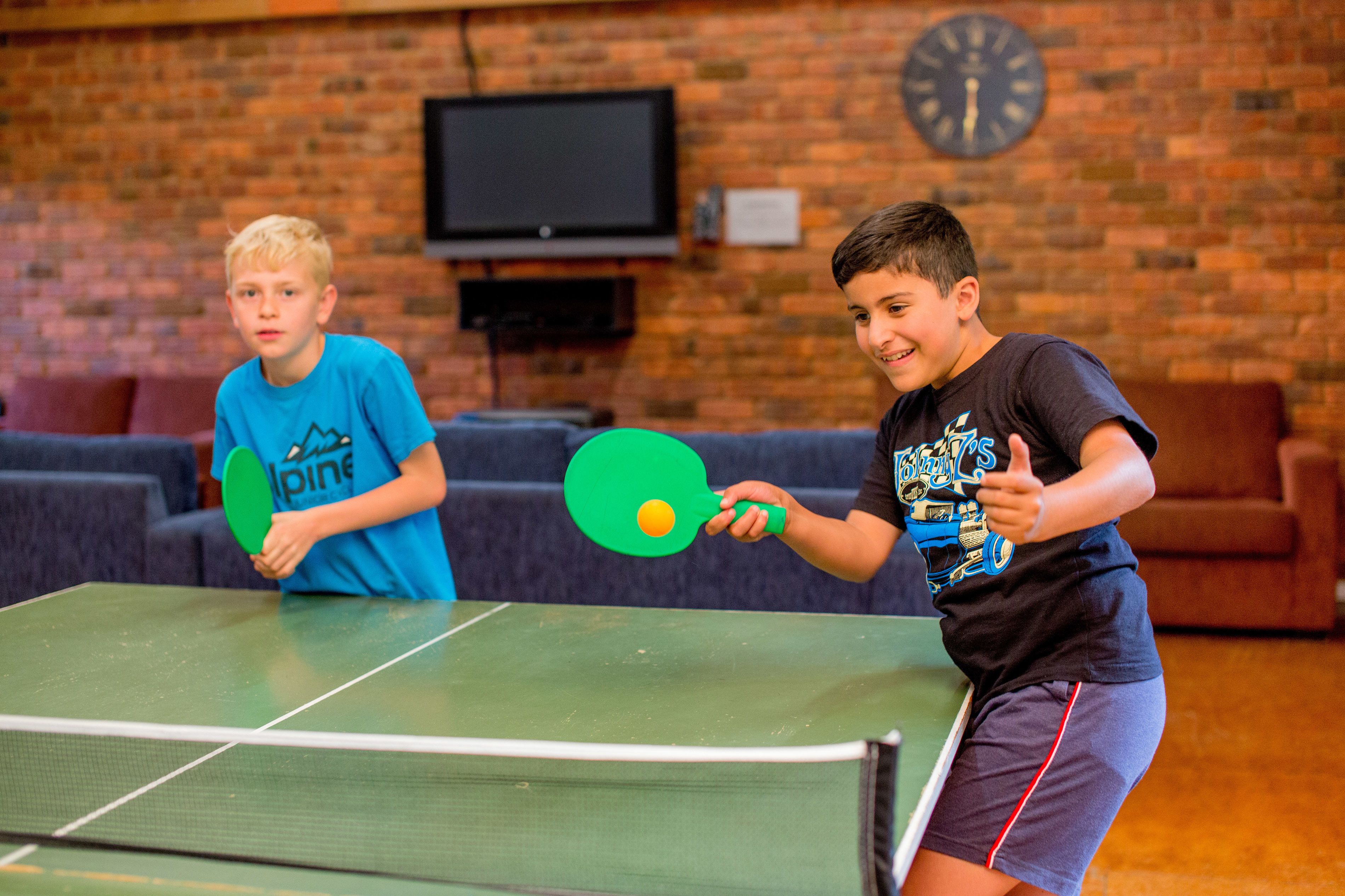 Two individuals playing table tennis in a room with a brick wall, a television, and a clock.