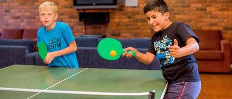 Two individuals playing table tennis in a room with a brick wall, a television, and a clock.