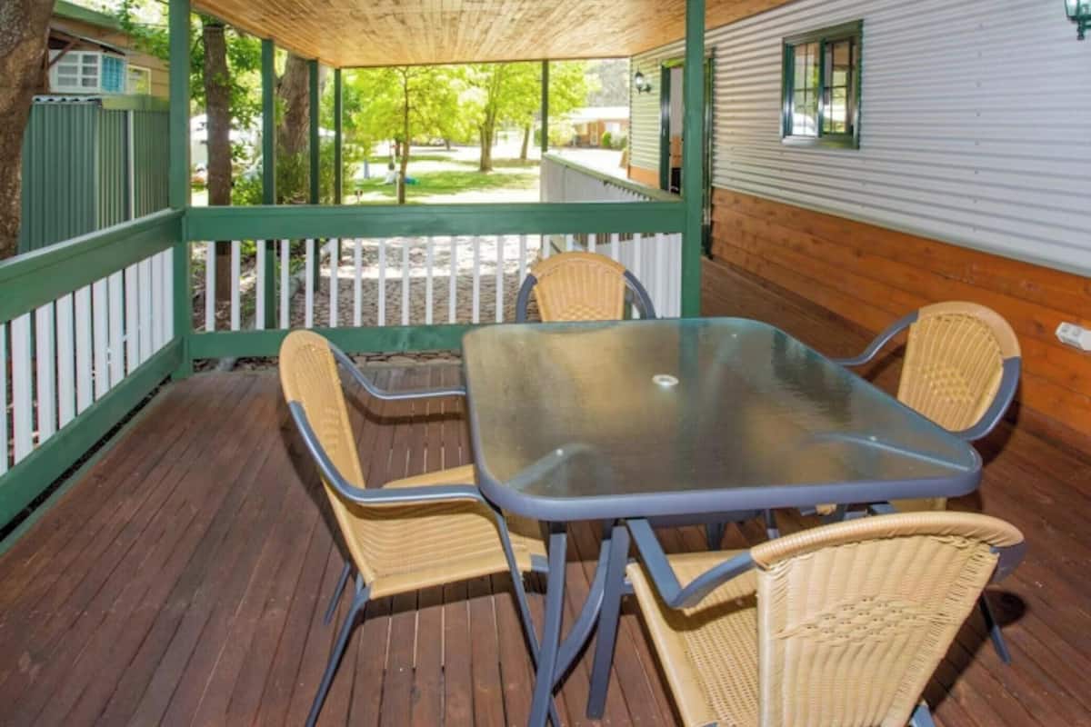 A covered outdoor seating area with a table and four chairs, surrounded by a wooden deck and a green railing.