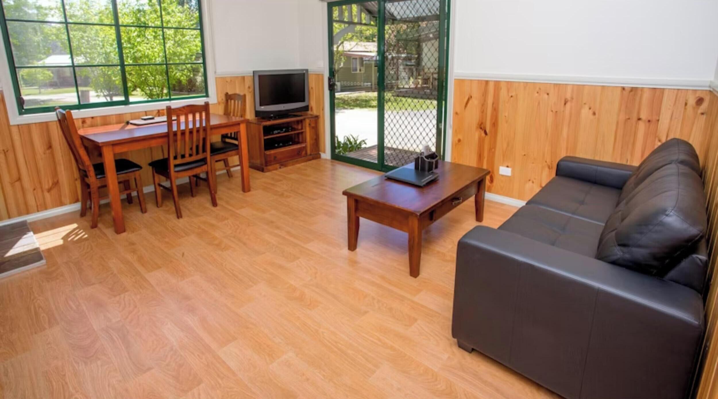 A furnished room with a wooden floor, a sofa, a TV, a dining table with chairs, and a sliding glass door leading outside.