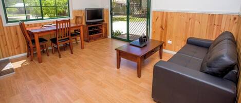 A furnished room with a wooden floor, a sofa, a TV, a dining table with chairs, and a sliding glass door leading outside.