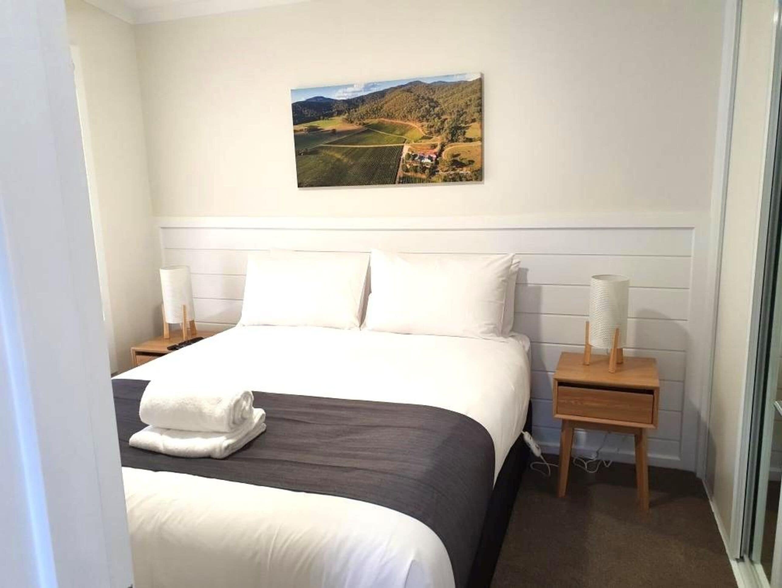Habitación de hotel con una cama grande, ropa de cama blanca, un cuadro de un paisaje y dos mesitas de noche con lámparas.