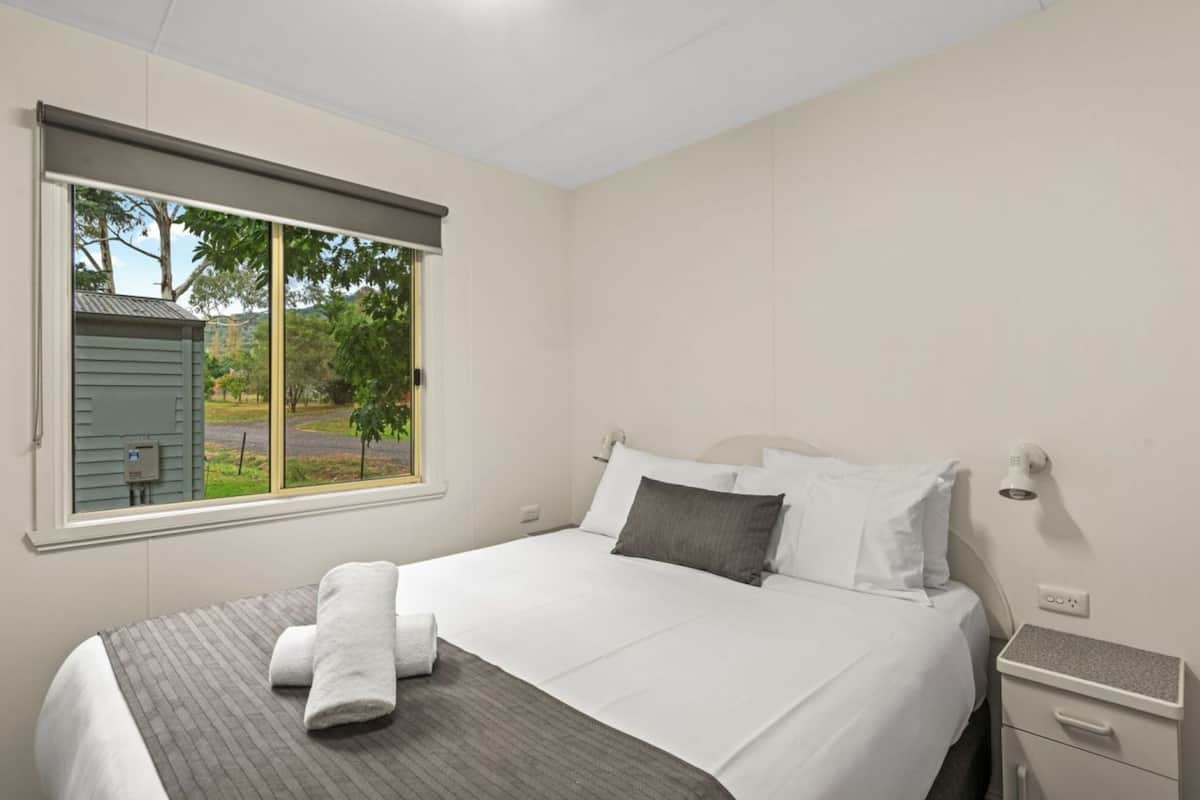 A hotel room with a bed, a nightstand, a lamp, a window with a view of trees and a building outside.