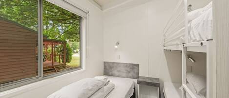 A bedroom with a bed, a desk, and a bunk bed. There is a window with a view of greenery outside.