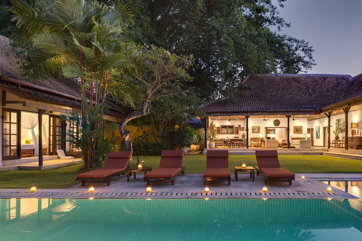 A poolside area with lounge chairs and candles, surrounded by a house with a tiled roof and a well-maintained lawn.